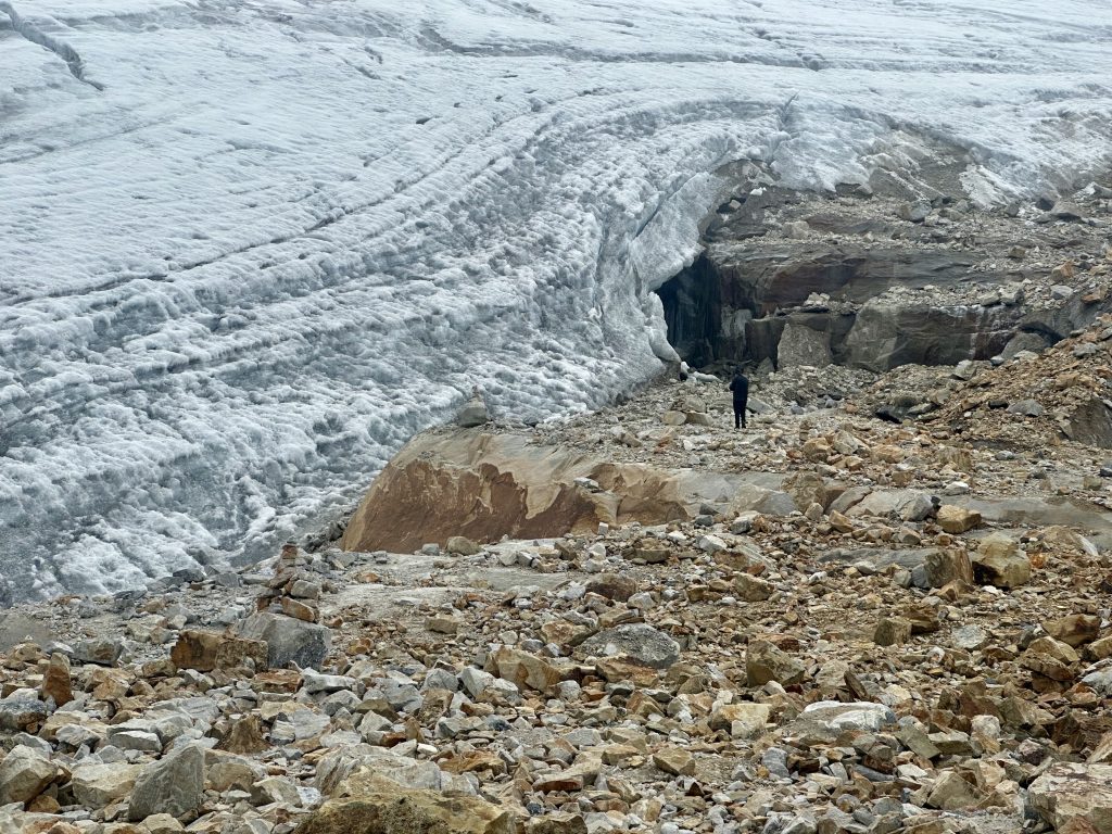 A lone figure stands before the retreating glacier in the Sierra Nevada del Cocuy, beneath a massive blue-white ice wall with a dark meltwater cave at its base, surrounded by large amber and grey boulders.