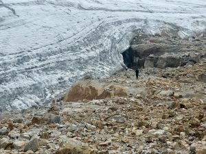 A lone figure stands before the retreating glacier in the Sierra Nevada del Cocuy, beneath a massive blue-white ice wall with a dark meltwater cave at its base, surrounded by large amber and grey boulders.