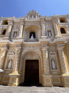 A close-up view of an ornate church facade featuring intricate white and yellow decoration. The building showcases detailed carvings, including columns and figures of saints, with a large arched entrance framed by decorative elements. Above the entrance, a statue is positioned in a niche, under a segment of ornate design, complemented by a clear blue sky in the background. The architecture reflects a blend of Baroque and colonial styles. - Santa Catalina church
