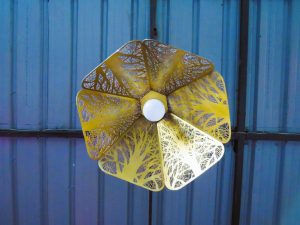 A gold hexagonal ceiling pendant lamp with laser-cut tree patterns and a white bulb is seen from below against a grey corrugated metal ceiling with a dark beam.