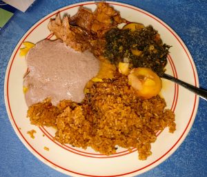 A white plate with red trim containing spiced rice, steamed greens, boiled potatoes, beef stew, chicken, and peanut sauce, set on a blue textured surface.