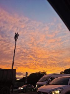 A vibrant sunset sky filled with orange and pink clouds above a busy roadside scene, with cars and silhouettes creating an urban evening atmosphere