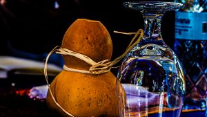 A clear wine glass filled with a golden-amber drink sits on a rustic wooden table, with a large dried brown calabash gourd behind it.