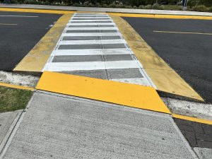 
A close-up view of a pedestrian crosswalk at a street corner. The crosswalk features white stripes and is bordered by a yellow ramp, which transitions from the sidewalk to the road