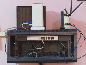 A front-facing shot of a black wall-mounted network rack containing a 16-port network switch with several connected Ethernet cables. Placed on top of the rack are a white wireless router, a black modem with dual antennas. The equipment is set against a light pink wall, with a tangle of black and grey cables visible inside 