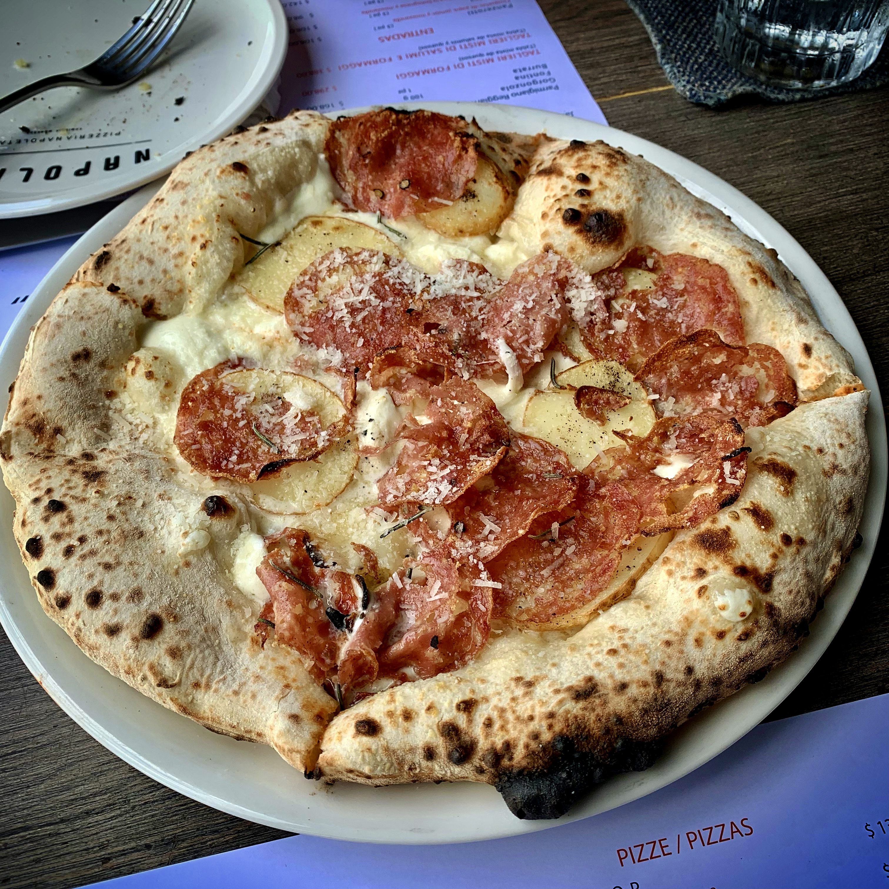 A Neapolitan-style pizza on a white plate, topped with salami, thin potato slices, fresh rosemary and grated Parmigiano on a white base, with a wide charred and blistered cornicione crust. The restaurant's Italian-Spanish menu is visible alongside on a wooden table.