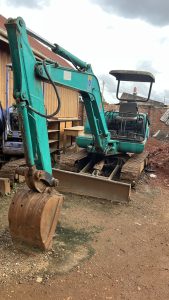 An excavator parked in a yard with another tractor and a roofed container in the background.