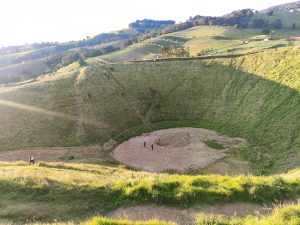 A large, grassy crater surrounded by rolling hills and trees, with several people standing at the bottom of the crater. Sunlight casts a warm glow on the landscape, highlighting the green slopes and the dry, cracked ground in the crater. There are paths leading down into the crater, and some people are seated or walking along the rim. Cráter la Olla, San Juan de Chicuá - Costa Rica
