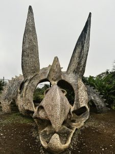 A large dinosaur skull sculpture with horns, set outdoors among greenery under a cloudy sky.