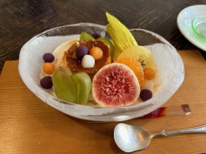 A beautifully arranged dessert in a clear oval bowl, featuring a variety of colorful fruits including grapes, figs, oranges, kiwi, and small round fruit garnishes.