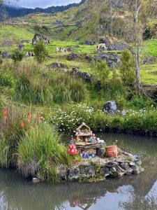 A small rock island in a highland pond near Güicán, Boyacá, holds a folk nativity scene with garden gnomes and ceramic figures, surrounded by flowers and grazing cattle on the hillside.