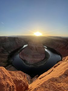Sunset at Horseshoe Bend, Page, Arizona. The sun is low on the horizon, casting a warm golden light over the layered rock formations and illuminating the landscape's textures.