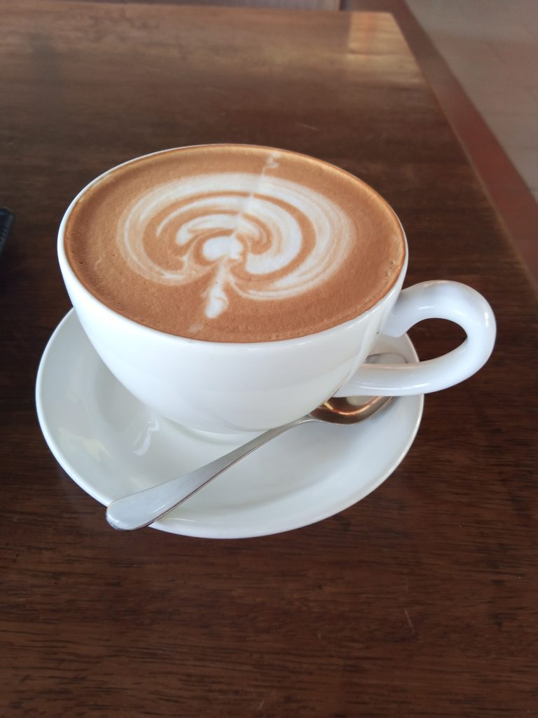 A white ceramic cup of coffee with circular latte art sits on a saucer with a small silver spoon on a dark wooden table, viewed from a slightly elevated angle.
