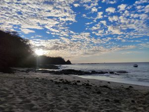 Beautiful coastal scene at dawn or dusk. A sandy beach with scattered rocks leads toward a calm ocean, where a small boat is visible in the distance. The sky is the focal point, featuring a striking pattern of altocumulus clouds illuminated by the soft light of the sun, which is partially hidden behind a dark, forested hillside.