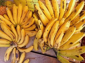 Several bunches of yellow bananas with small brown spots arranged on a wooden surface, with a partially visible pineapple and a yellow mesh bag in a sunlit market setting.