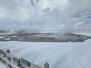 A winter scene at a geothermal area features the Morning Geyser venting steam into the cloudy sky. A wooden fence with signs for “Morning Geyser” and “Fountain Geyser” is visible in the foreground, while patches of snow cover the ground. The landscape is dominated by rocky terrain and geothermal features, with a backdrop of distant mountains shrouded in mist.