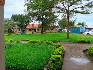 Landscaped garden with trimmed bushes and a tiled-roof building, with vehicles and trees under a cloudy sky.