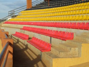 Rows of plastic stadium seats in tiers of black, yellow, and red are mounted on concrete steps, with a metal railing along a staircase.