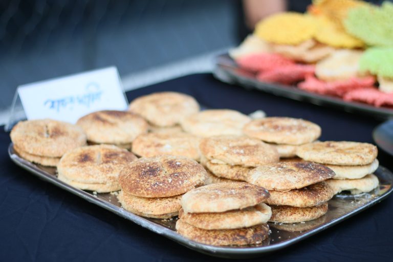 A metal tray filled with round, golden-brown baked goods, each with a slightly textured top and a sprinkling of sesame seeds. In the background, there is a colorful assortment of other snacks, and a small sign in front of the tray indicating the name of the food. The setting includes a black tablecloth and a blurred background.