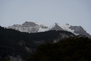 Snow-covered mountains in Switzerland with forested hills in the foreground and cloudy sky.