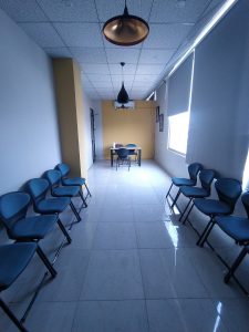 A narrow hallway features several rows of blue chairs on either side, leading toward a small table and two chairs at the far end. The room has beige and gray walls, with large windows covered by roller shades allowing natural light to enter. 