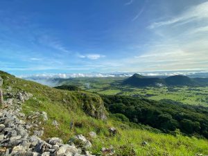 A scenic view from a mountain ridge overlooking a lush green valley. The foreground features rocky terrain and grass, while the background is dominated by rolling hills and low-lying clouds. A bright blue sky with a few wispy clouds stretches overhead, creating a peaceful and natural atmosphere.