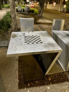 A concrete table designed for outdoor use features a checkered pattern on its top, suitable for chess. Two simple, matching concrete seats are positioned beside the table. The surrounding environment includes a path lined with trees and low shrubs, with a few scattered leaves on the ground. In the background, there are faint hints.