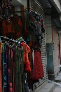 A street view displaying a variety of colorful clothing items hanging for sale. The garments include patterned shawls, skirts, and trousers with intricate designs, set against a backdrop of closed shop shutters. The scene reflects a vibrant marketplace atmosphere.