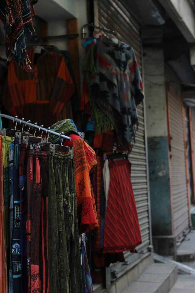 A street view displaying a variety of colorful clothing items hanging for sale. The garments include patterned shawls, skirts, and trousers with intricate designs, set against a backdrop of closed shop shutters. The scene reflects a vibrant marketplace atmosphere.