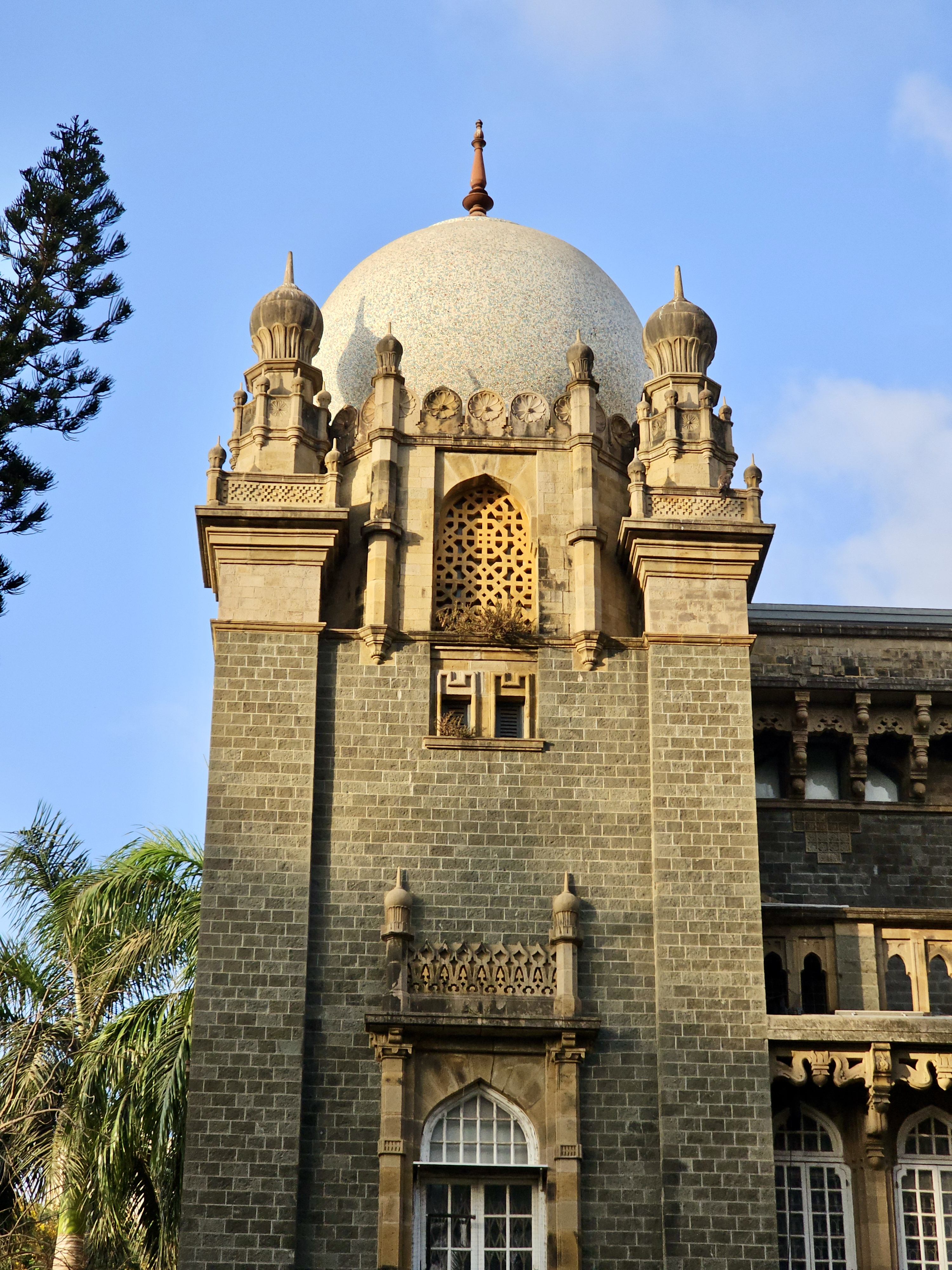 A historic building portion with a dome and intricate stonework. Sunlight highlights the textures, giving the structure a grand and elegant appearance. It is part of the Chhatrapati Shivaji Maharaj Vastu Sangrahalaya in Mumbai.