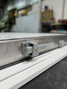 A close-up view of a metallic locking mechanism attached to a sliding door or window frame, with a white base underneath. 