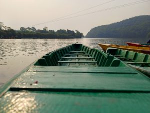 A view from inside a green wooden boat floating on a calm lake, with forested hills on both sides and a hazy sky above.