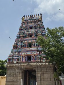 Colorful South Indian temple tower (gopuram) adorned with intricate sculptures of deities and figures, rising above a stone entrance, with trees on the side and birds flying in a partly cloudy sky.
