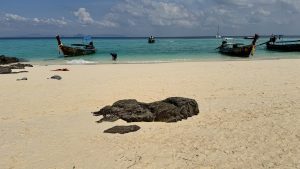 A wide beach scene shows clear turquoise water and several wooden longtail boats near the shore. In the foreground, a dark jagged rock sits in the middle of the light tan sand. A person is seen near one of the boats under a sky with white clouds.