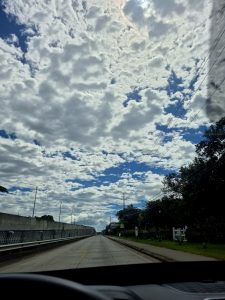 A long open road under a dramatic sky filled with thick white clouds and bright blue patches, creating a peaceful and cinematic daytime driving scene