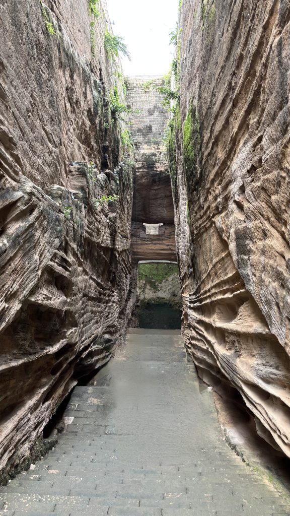 A narrow stone path goes down between tall rocky walls. Steps lead to a small pool of water below. Above, a stone bridge crosses the gap, with some green plants growing on the wet rocks.