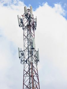 A tall communication tower with multiple antennas and satellite dishes mounted at the top, set against a backdrop of a partially cloudy blue sky.