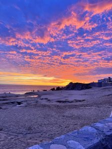 A colorful sunset fills the sky over the coast of Los Cabos, Mexico, with shades of pink, purple, and orange reflecting above the beach.