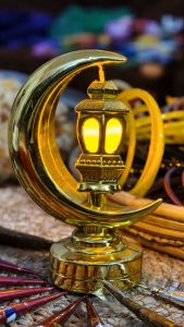 A gold crescent moon and lantern ornament glowing warmly on a woven surface, with henna tubes nearby and a festive blurred background.