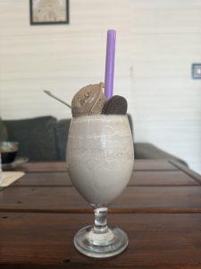 A creamy milkshake in a clear glass, topped with a scoop of brown ice cream, a purple straw, and an Oreo cookie on a wooden table.