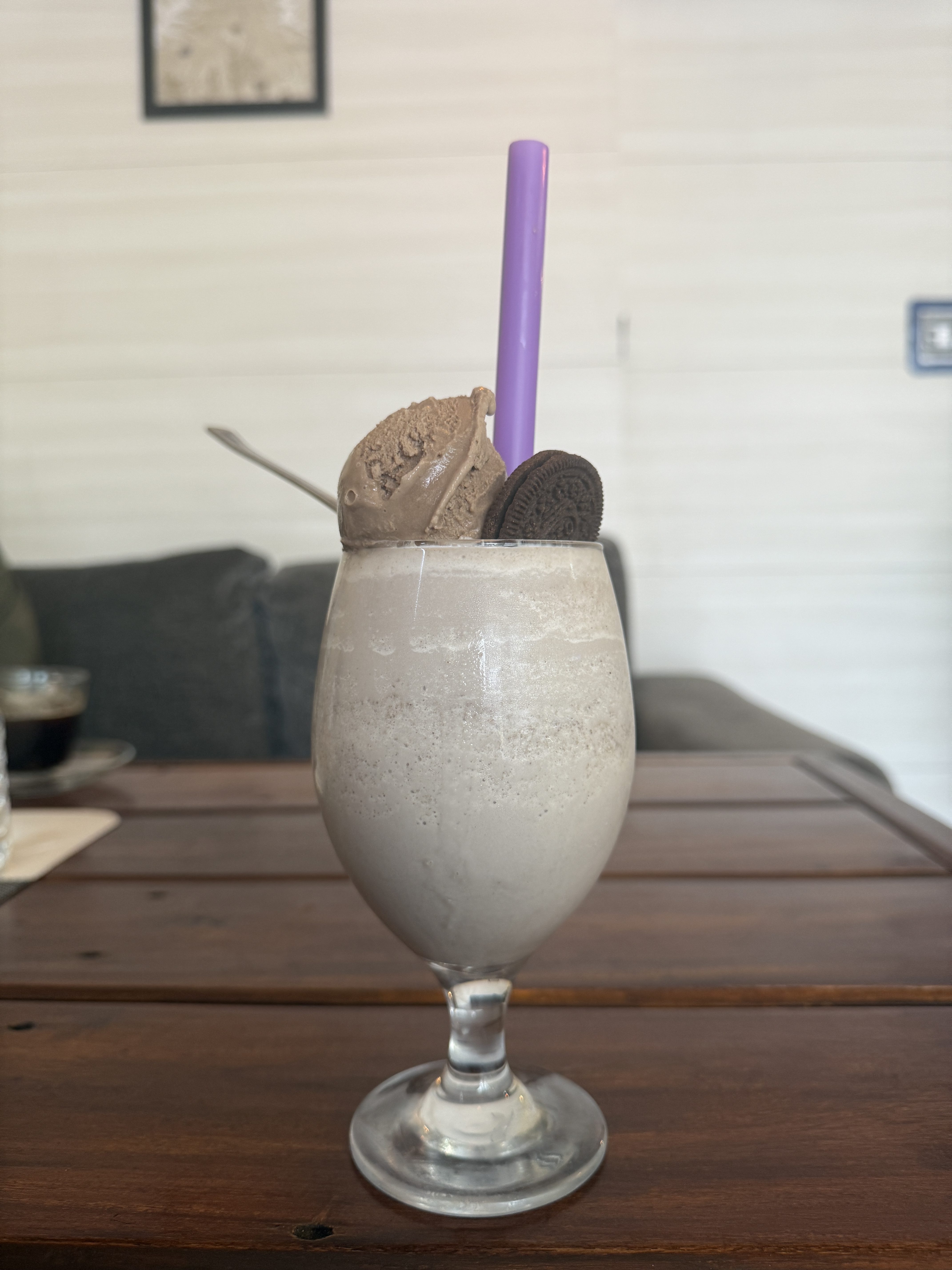 A creamy milkshake in a clear glass, topped with a scoop of brown ice cream, a purple straw, and an Oreo cookie on a wooden table.