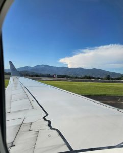 A perspective from inside a plane looking out over the wing towards a grassy runway and a beautiful range of blue mountains.