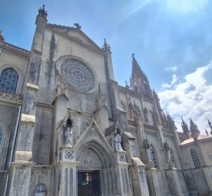 A close-up view of a Gothic-style church facade, featuring a large round stained glass window and intricately designed pointed arches. The building is adorned with statues and decorative elements, set against a bright blue sky with a few fluffy clouds. The structure shows signs of weathering, adding to its historical character.