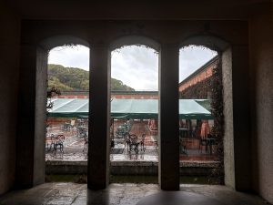 A view through three arched windows showing a rainy outdoor seating area. Outside are metal tables and chairs under green canopies. The scene is in Okayama’s Ivy Square.