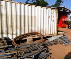 A large white shipping container sits in a dusty yard, surrounded by piles of blue metal scaffolding poles and thick, rusted industrial hoses. In the background, a red container structure and a yellow forklift are visible.