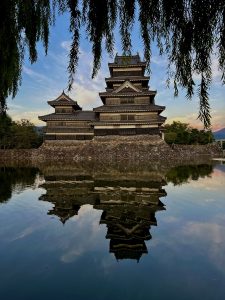 Matsumoto Castle rises above its moat at dusk, framed by willow branches and reflected in the still water.
