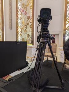 A professional video camera mounted on a tripod is positioned on a black carpeted area, with a black backdrop and chairs in the background. 
