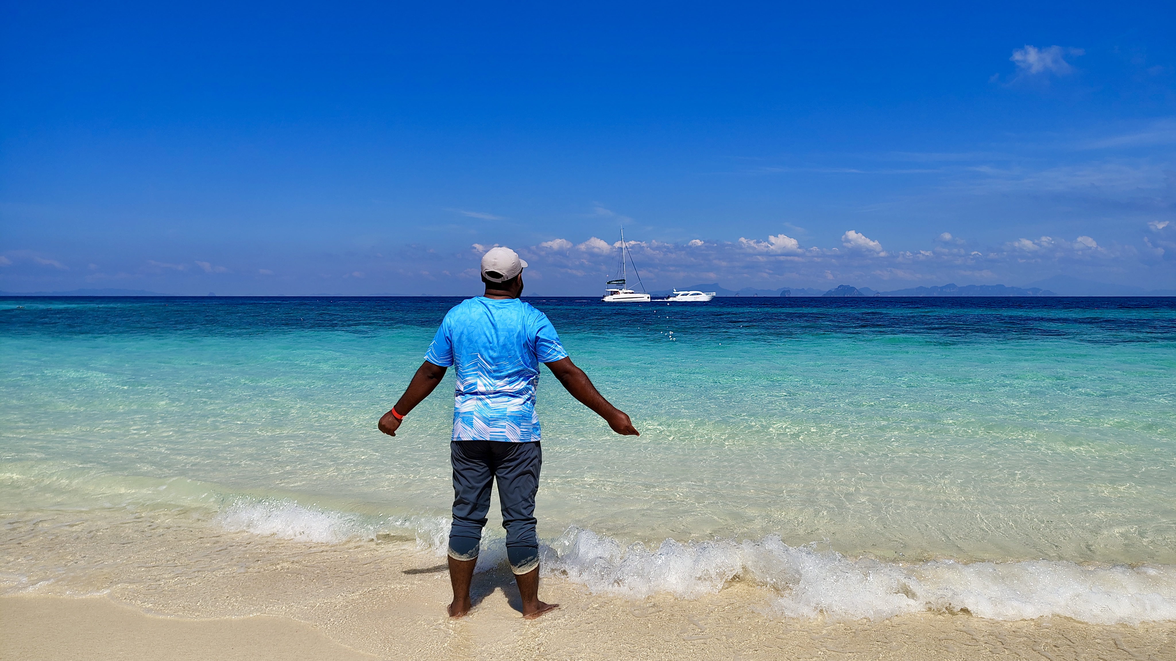 A man in a blue shirt and cap stands by the ocean with arms slightly out, facing the sea under a bright sky.