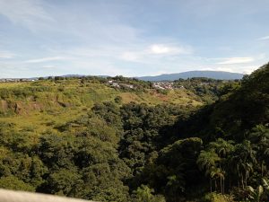 Panoramic view of lush green hills and dense forest, with a small settlement in the distance. Rolling terrain, distant mountains, and a clear blue sky create a calm, natural landscape in Alajuela.