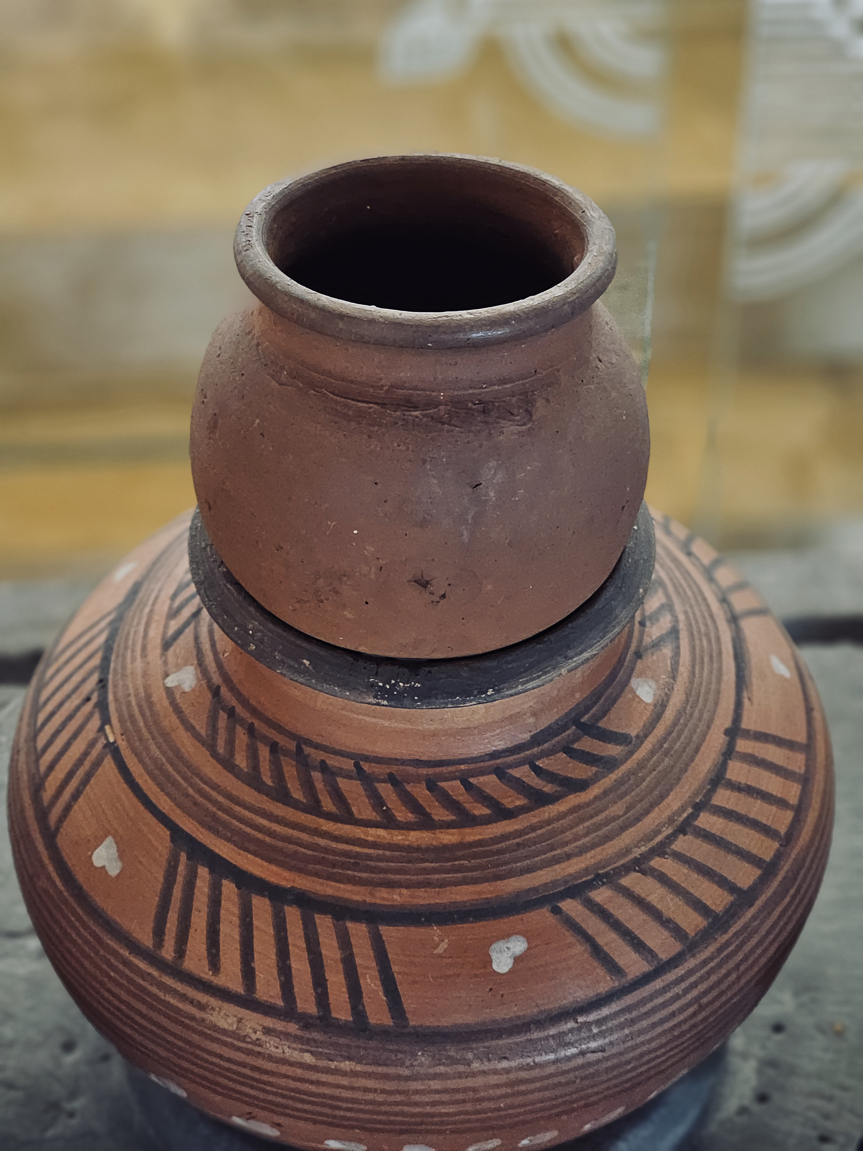 A handcrafted terracotta pot with black geometric patterns and small white accents. A smaller plain clay pot sits on top, creating a simple stacked look. Shot indoors in Andheri, Mumbai.